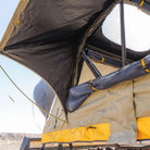 Open CVT rooftop tent with yellow trim, revealing the interior support poles—including the corrosion-resistant Pioneer Spring Rod—and a clear skylight window, set against a bright sky and rocky landscape in the background.