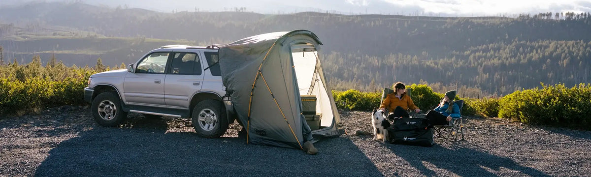 An SUV with a tent attached is parked on gravel; two people sit in chairs nearby with a dog, surrounded by mountains and greenery under a cloudy sky.