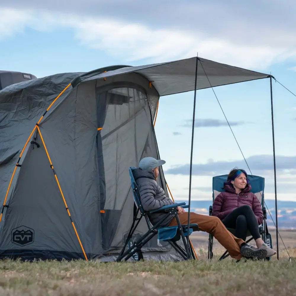 Two people relax in camping chairs under an awning attached to a spacious gray tent, set up on grassy ground with a scenic sky and distant hills in the background—the ideal arrangement for car camping or overlanding adventures, especially with the convenience of the CVT Timber Creek SUV Annex close at hand.
