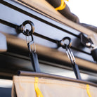 Close-up of two black metal hooks attached to a rail, each supporting a strap connected to a beige fabric item with yellow trim—the CVT Mt. Hood/La Sal Shoe Bag—designed for convenient rooftop tent shoe storage as part of outdoor or camping gear.