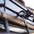 Close-up of a black metal ring and carabiner fastened to the rail of a roof rack with secured straps on a vehicle. Perfect for attaching accessories such as the CVT Mt. Hood/La Sal Shoe Bag or other rooftop tent shoe storage solutions, this setup keeps your essentials organized while traveling. The background is softly blurred.