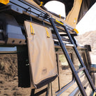 A beige Mt. Hood/La Sal Shoe Bag by CVT, featuring yellow straps, is attached to a black roof rack ladder on an off-road vehicle parked near rocky terrain.