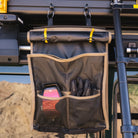 A CVT Mt. Hood/La Sal Shoe Bag, featuring two pockets holding gloves and a red-black device, is attached to a metal roof rack on a vehicle, serving as an ideal rooftop tent shoe storage solution against the backdrop of a mountainous landscape.