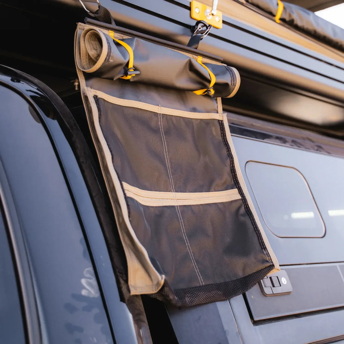 A black and tan CVT Mt. Hood/La Sal Shoe Bag hanging storage organizer is attached to the side of a vehicle with camping gear on the roof. Designed as a rooftop tent shoe storage solution, this organizer features mesh and zippered pockets, making it well-suited for outdoor or travel use.