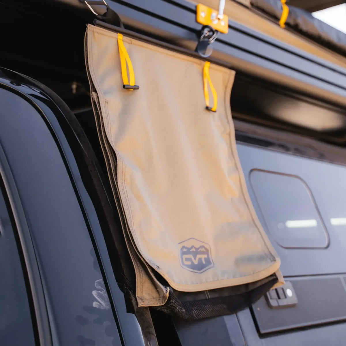 A Tan Mt. Hood/La Sal Shoe Bag from CVT with yellow handles hangs from a black vehicle’s roof rack. Designed as a rooftop tent shoe storage solution, the bag features a mountain logo and is attached near a window, underneath a mounted awning.