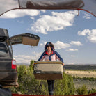 A woman in sunglasses carries camping gear, including the CVT Soft Side Storage™ bag, near an open car trunk in a scenic outdoor setting featuring trees, grass, and a blue sky with clouds.