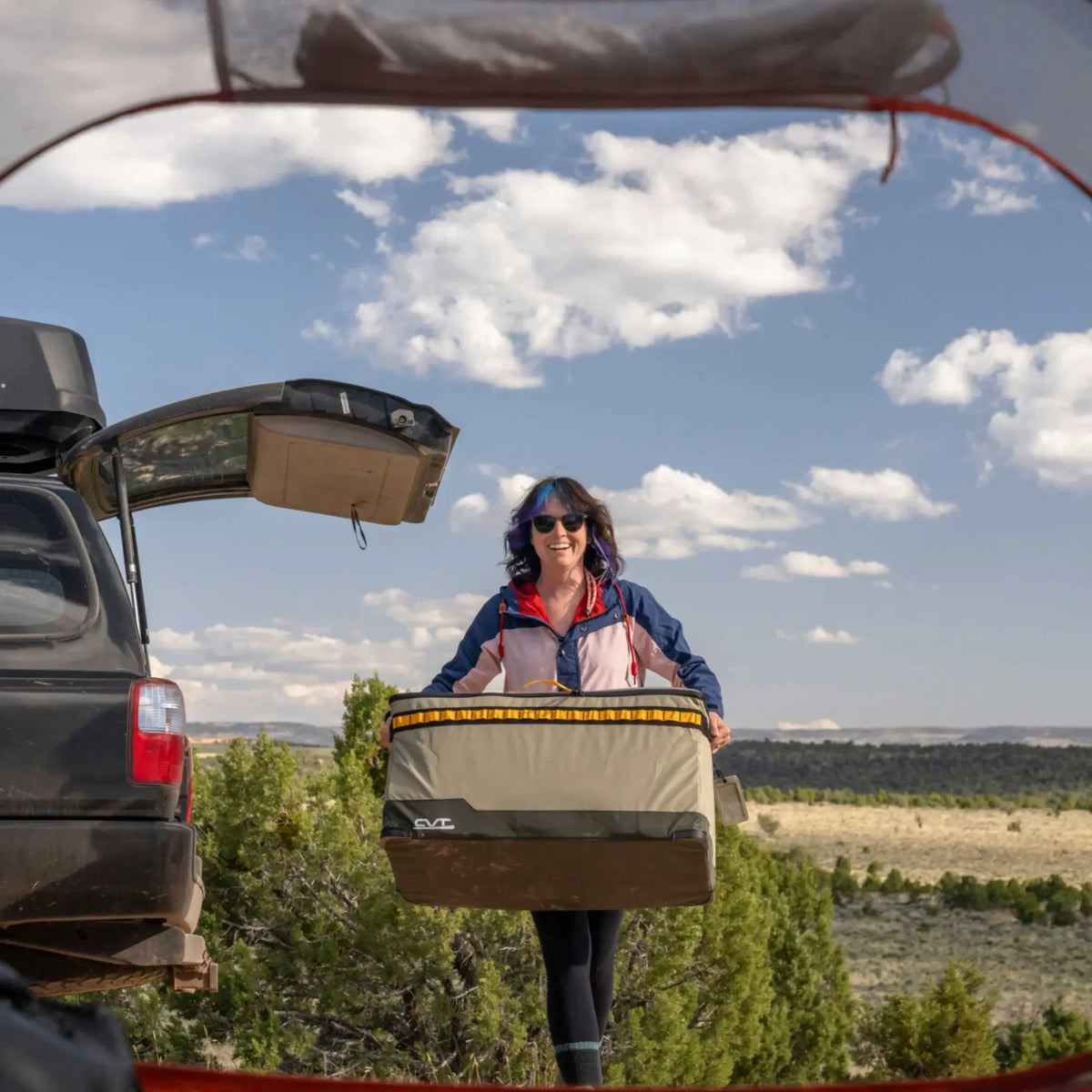 A woman in sunglasses carries camping gear, including the CVT Soft Side Storage™ bag, near an open car trunk in a scenic outdoor setting featuring trees, grass, and a blue sky with clouds.