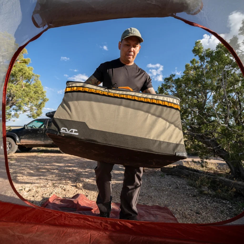 A person stands outside a tent, holding a large, rectangular storage container. There is a car and trees in the background under a sunny sky.