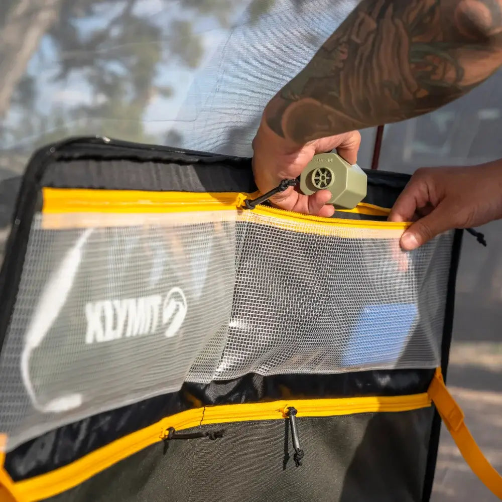 A person with tattooed arms places a small fan inside a yellow and mesh water-resistant Soft Side Storage™ pocket by CVT, attached to camping gear outdoors.