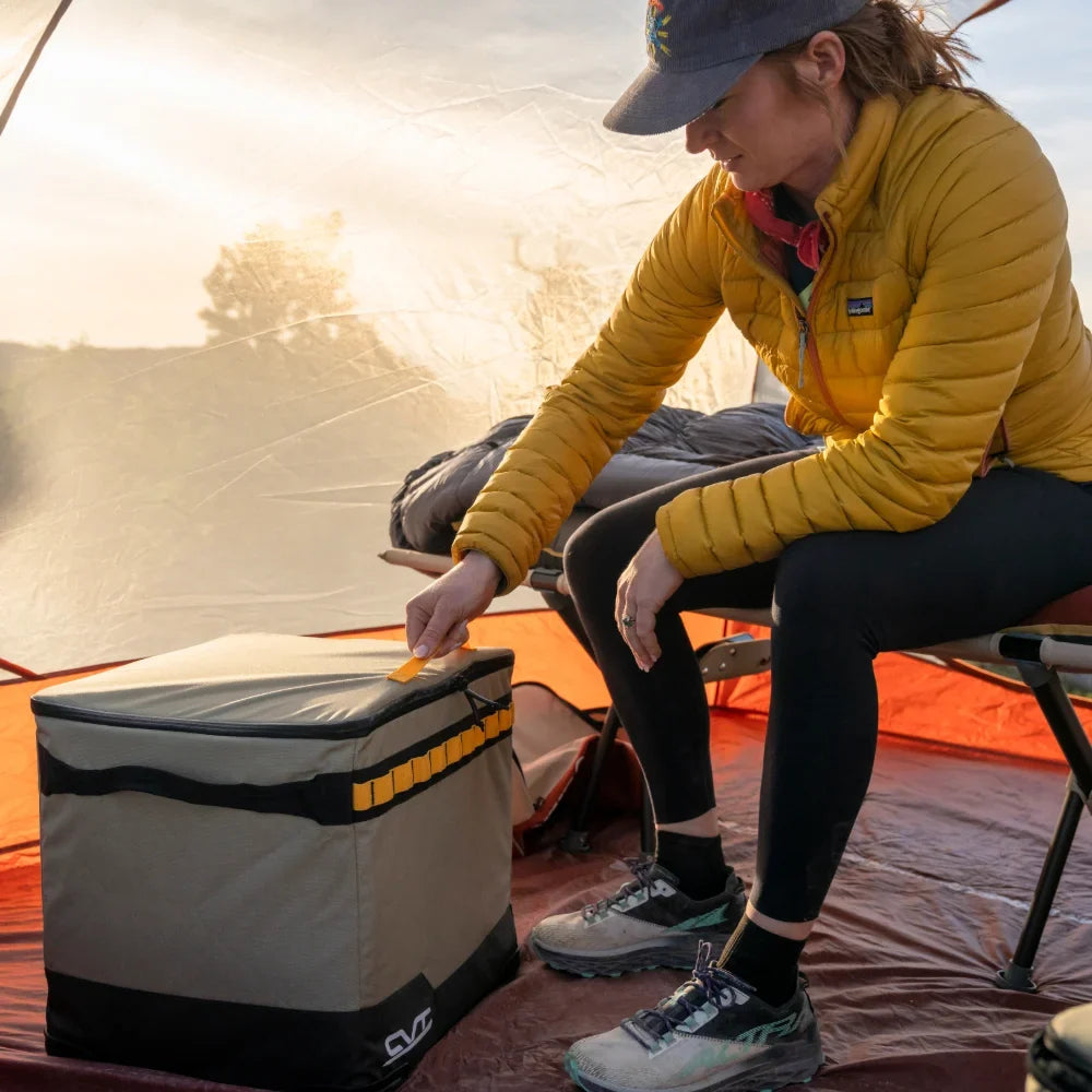 A person in a yellow jacket sits inside a tent, reaching for the CVT Soft Side Storage™ cooler—renowned for its water-resistant storage—resting on the ground beside a camping cot.