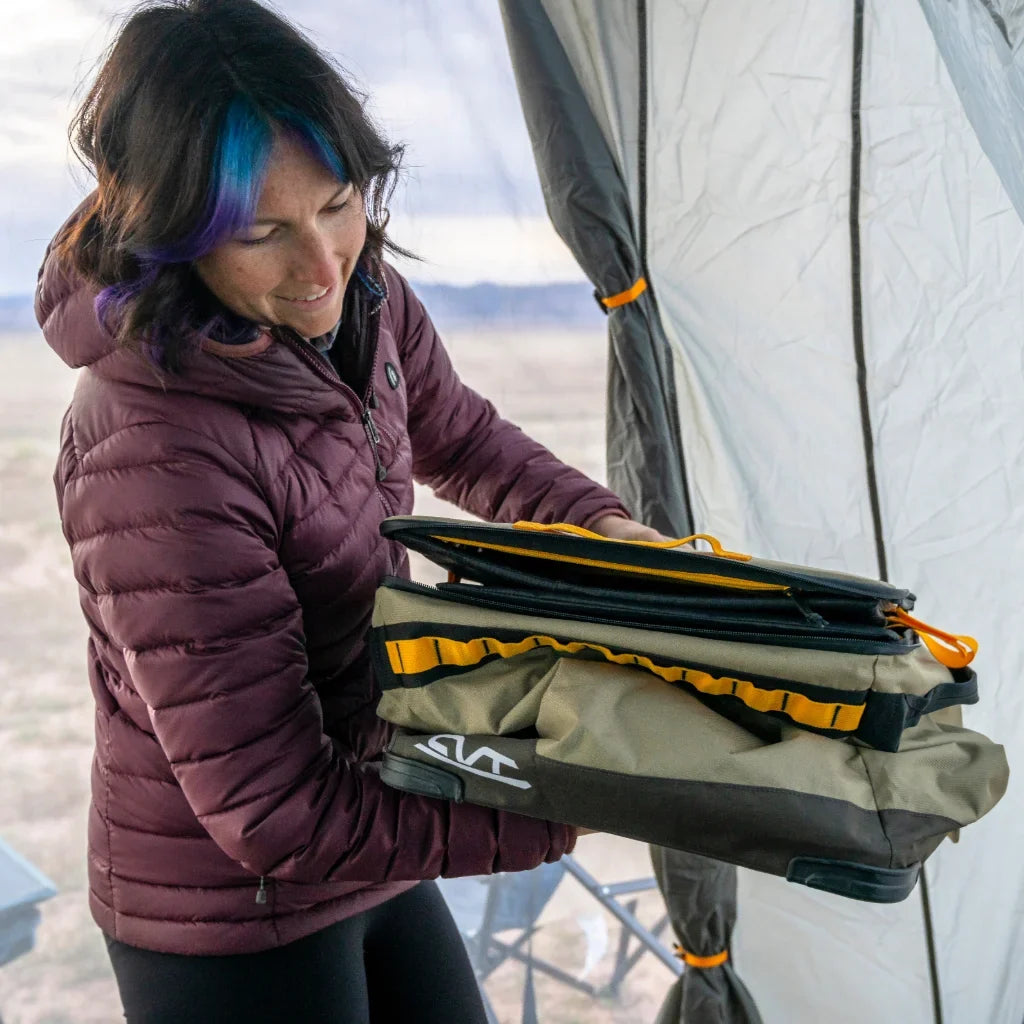 A person in a maroon jacket stands inside a tent, holding folded camping gear with yellow straps and a gray cover.