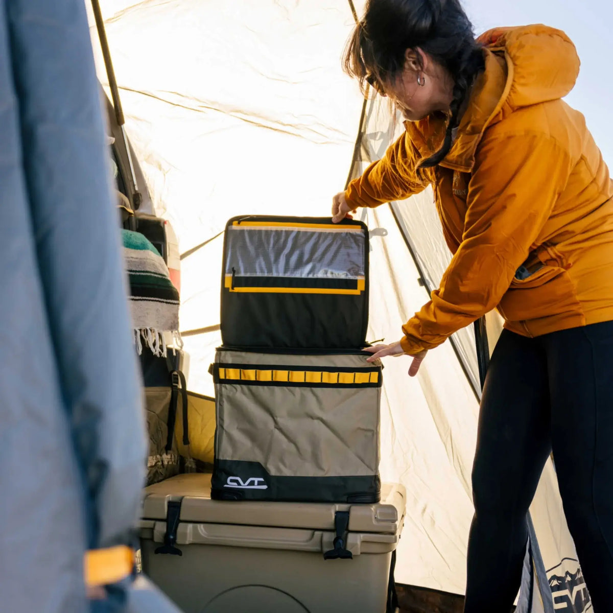 Wearing an orange jacket, a person stacks CVT Soft Side Storage™ bins—rectangular and collapsible—inside a tent, where sunlight filters in to highlight camping gear, water-resistant storage coolers, and blankets.