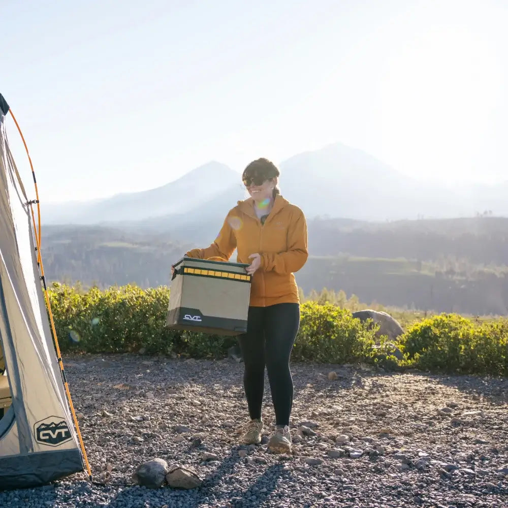 A person in an orange jacket carries the CVT Soft Side Storage™ box near a tent, with mountains and trees in the background under bright sunlight.
