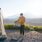 A person in an orange jacket carries the CVT Soft Side Storage™ box near a tent, with mountains and trees in the background under bright sunlight.