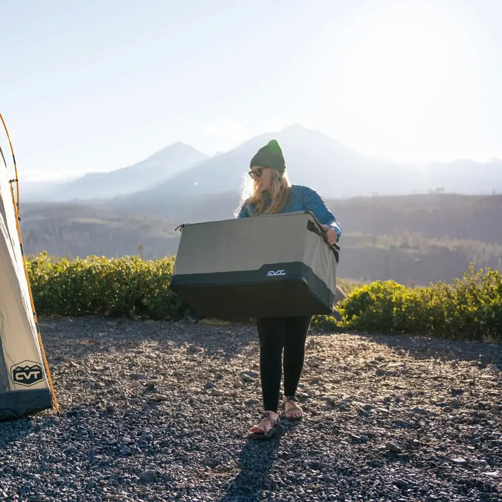 A person wearing a green beanie carries a large gray CVT Soft Side Storage™ container outdoors near mountains, standing on a gravel surface with sunlight in the background.