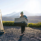 A person wearing a green beanie carries a large gray CVT Soft Side Storage™ container outdoors near mountains, standing on a gravel surface with sunlight in the background.