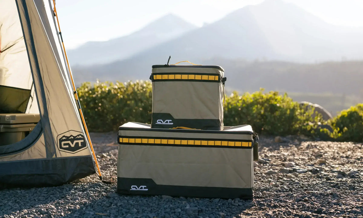 Two stacked, rectangular camping storage bins with yellow accents sit on rocky ground near a tent, with greenery and hazy mountains in the background.