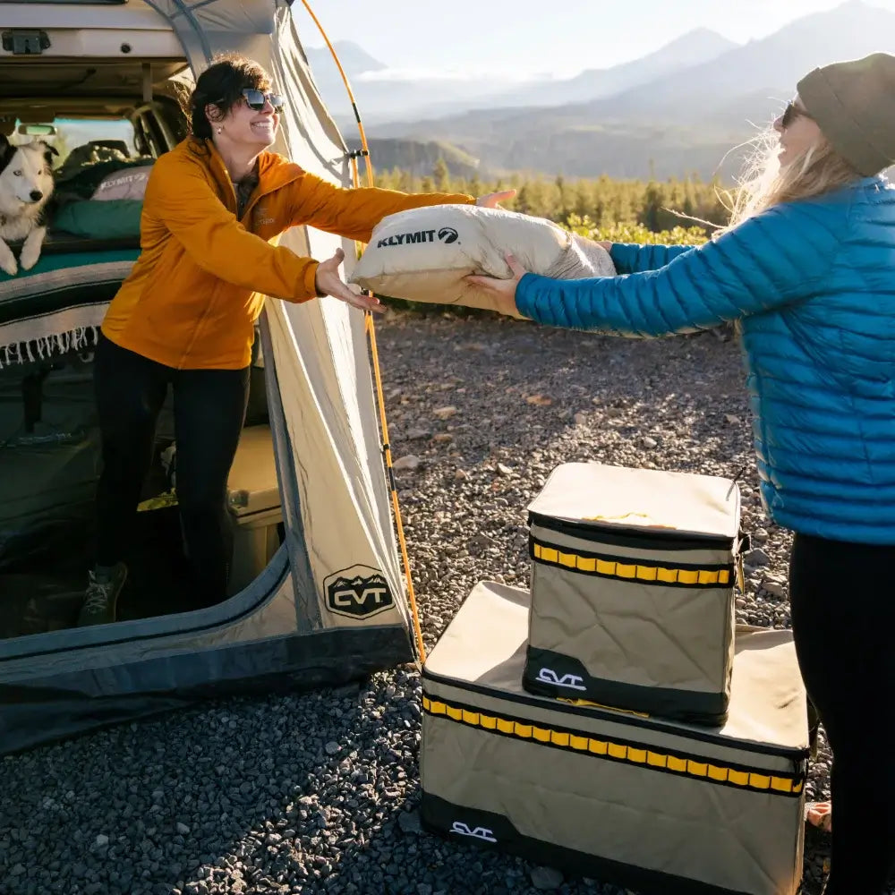 Two people exchange a Klymit sleeping pad outside a tent with a dog inside, surrounded by camping gear and mountains beneath clear skies. Nearby, CVT’s Soft Side Storage™ keeps their essentials protected and water-resistant for the adventure ahead.
