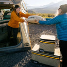 Two people exchange a Klymit sleeping pad outside a tent with a dog inside, surrounded by camping gear and mountains beneath clear skies. Nearby, CVT’s Soft Side Storage™ keeps their essentials protected and water-resistant for the adventure ahead.