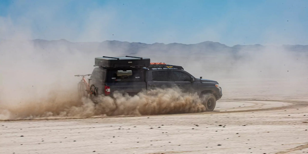 A dark pickup truck kicks up dust while driving fast across a dry desert landscape, with mountains visible in the background under a blue sky.