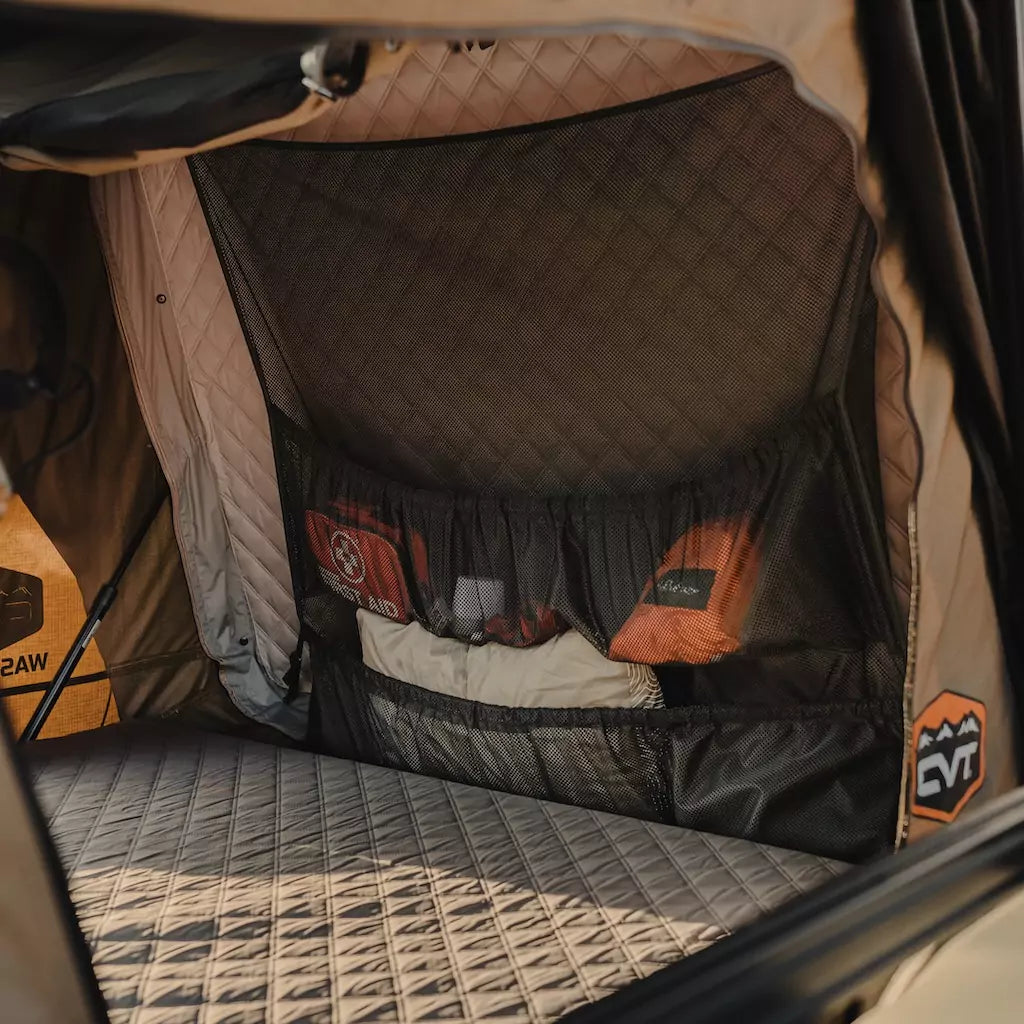 The image shows the interior of a tent with quilted walls and base. Mesh pockets hold various camping gear, including a first aid kit, inside the tent. Orange and black logo patches are visible on the walls.