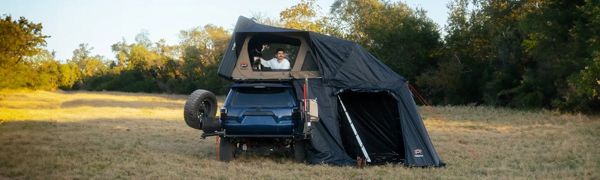 A man sits inside a rooftop tent mounted on a dark SUV, parked on dry grass near trees. The tent has an attached annex, and the scene is set during daylight in a natural outdoor area.