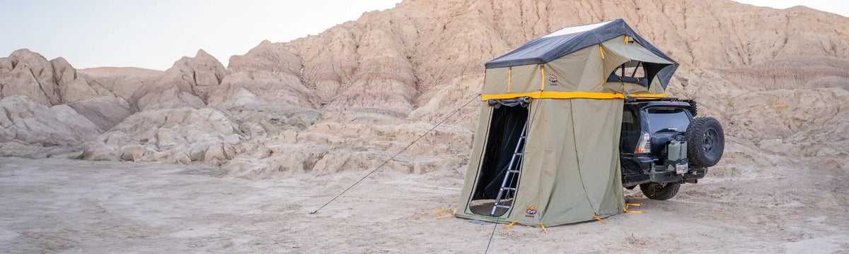 A rooftop tent with an annex is set up on a vehicle parked in a rocky desert landscape with beige, rugged hills in the background.