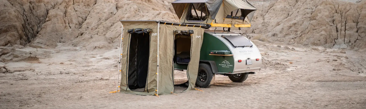 A green teardrop camper trailer with an attached tent, rooftop tent, and awning stands in a rocky desert landscape.
