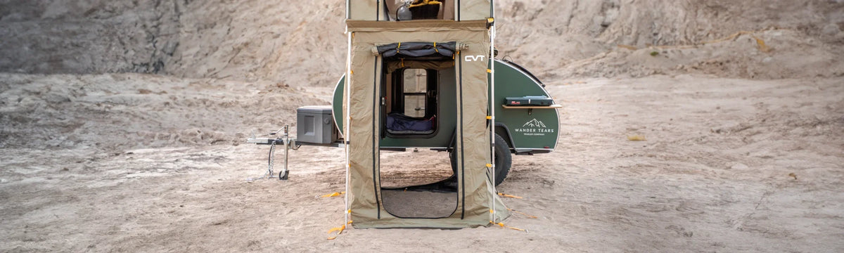 A tan rooftop tent with an attached ground enclosure stands next to a green teardrop camper in a barren, rocky desert landscape.