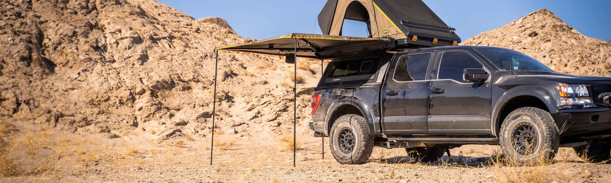 A black pickup truck with a rooftop tent and side awning is parked in a desert with rocky hills in the background under a clear blue sky.