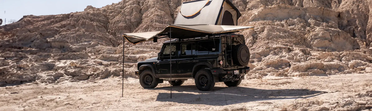 A black off-road vehicle with a rooftop tent and awning is parked on sandy terrain, surrounded by rocky hills under a clear sky.