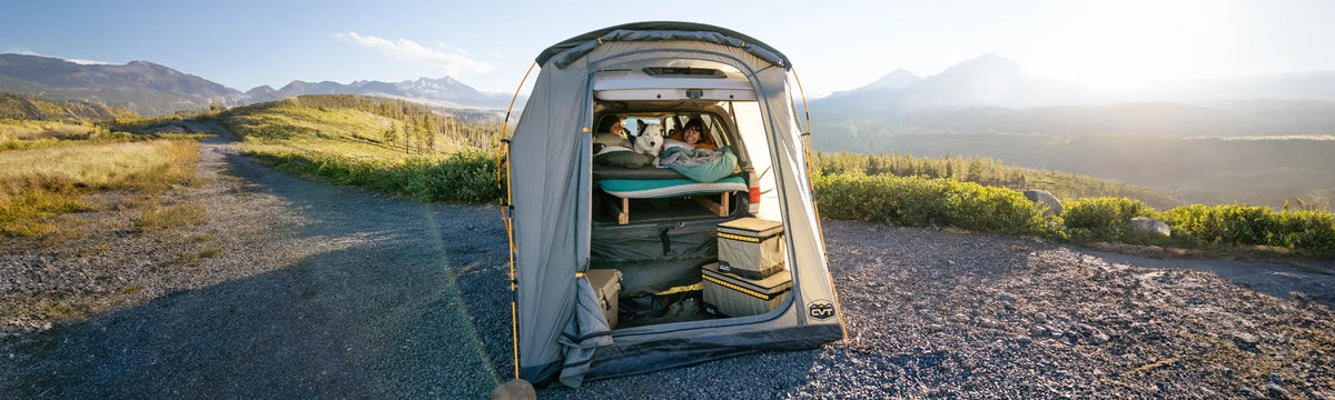A person and a dog relax inside a car outfitted with a bed and storage bins, parked in a tent with scenic mountains, grassy fields, and a bright sky in the background.