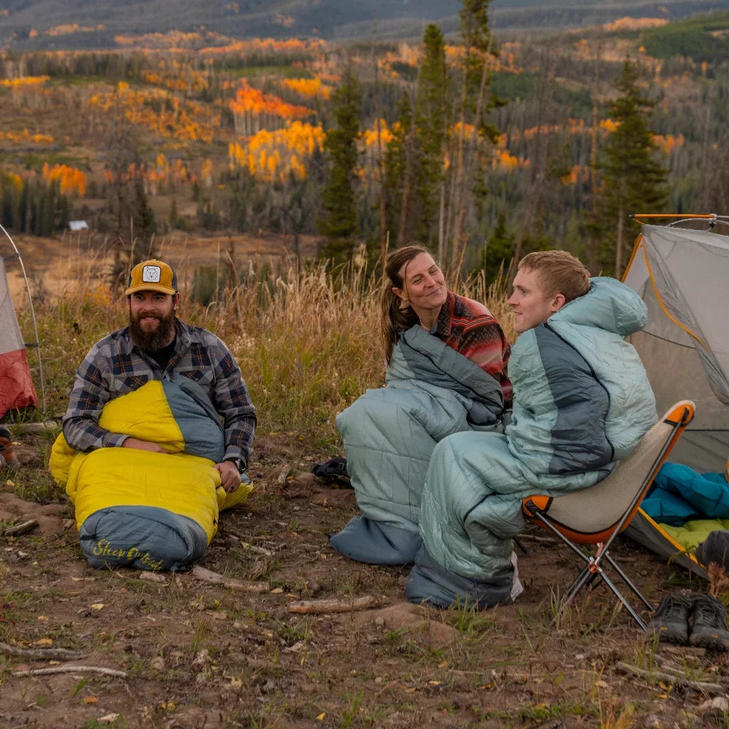 Three people sit outdoors in sleeping bags near a tent, surrounded by autumn trees and mountains. They appear to be camping, talking, and enjoying the scenic landscape in the cool weather.