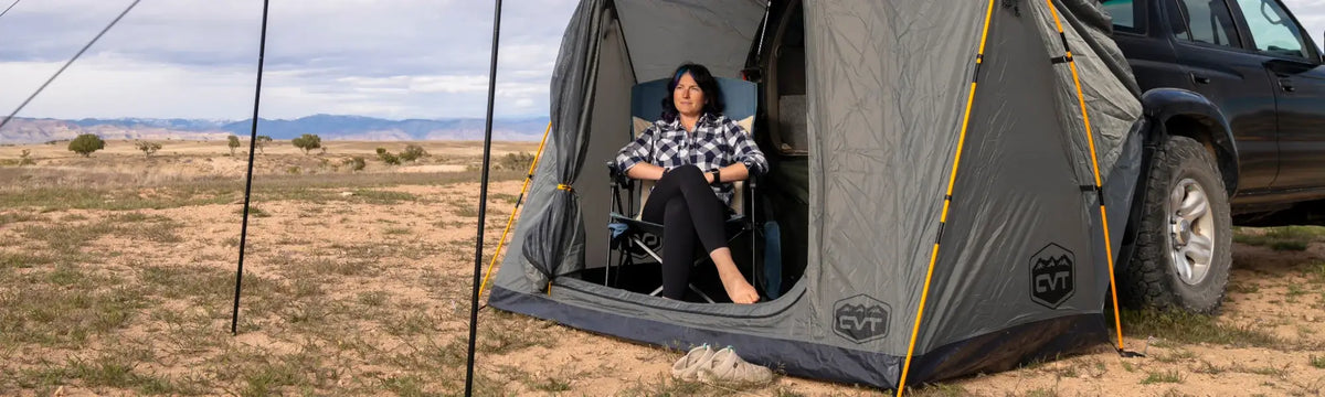 A person sits in a camping chair inside a tent attached to a black SUV, set up in a dry, open landscape with distant mountains under a cloudy sky.