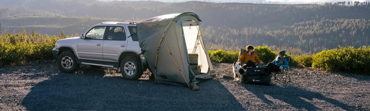 A silver SUV with a tent attachment is parked on gravel. Two people sit in camping chairs by the tent, talking, with gear and a dog nearby. Mountains and trees are visible in the background under a clear sky.