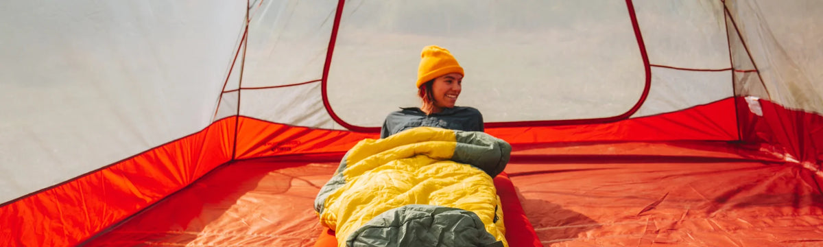 A person in a yellow beanie smiles while sitting in a yellow sleeping bag inside a red and white tent, with soft natural light coming in.