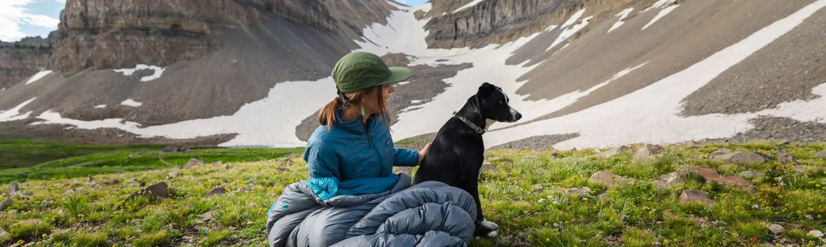 A person in a blue jacket and green cap sits on grass next to a black dog, surrounded by rocky mountains with patches of snow.