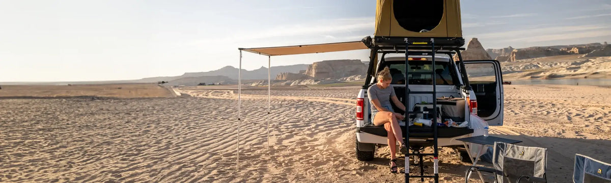 A person sits on the back of a vehicle with a rooftop tent in a desert landscape, working on a laptop, surrounded by camping gear, chairs, and a distant mountain range.