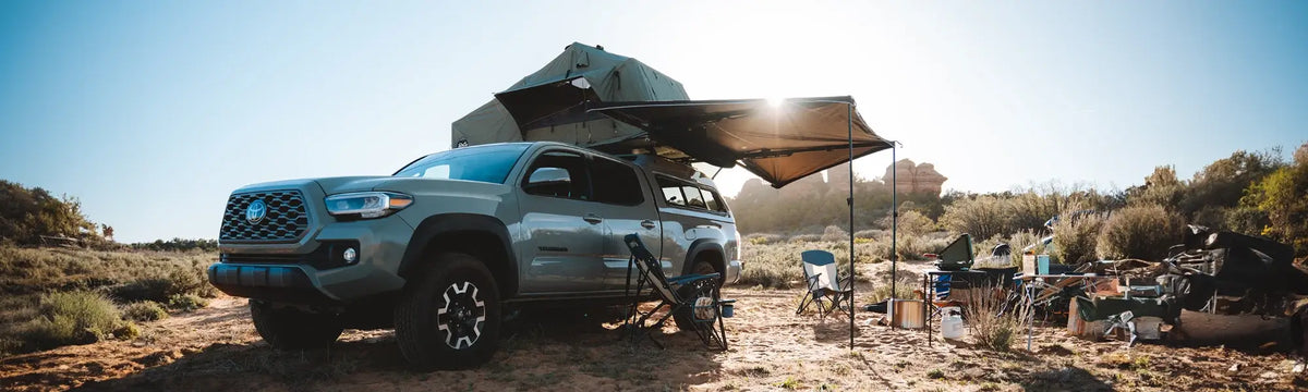 A gray pickup truck with a rooftop tent and awnings is set up for camping in a sunny, grassy area with trees, chairs, and supplies scattered nearby.