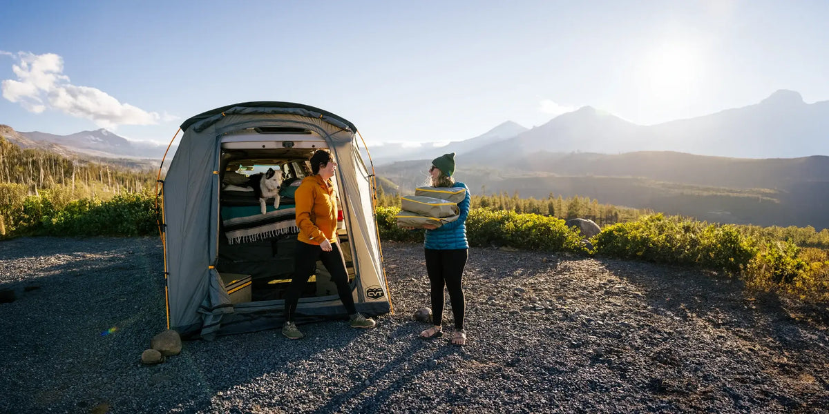 Two people stand next to a van with an attached tent in a scenic mountain area. One hands bedding to the other. A dog is inside the tent. The scene is sunny with mountains and trees in the background.