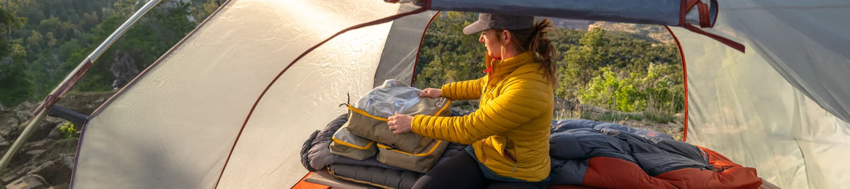 Person in a yellow jacket sits inside a tent, packing or unpacking a bag, with sleeping gear around and a scenic outdoor view seen through the tents open sides.