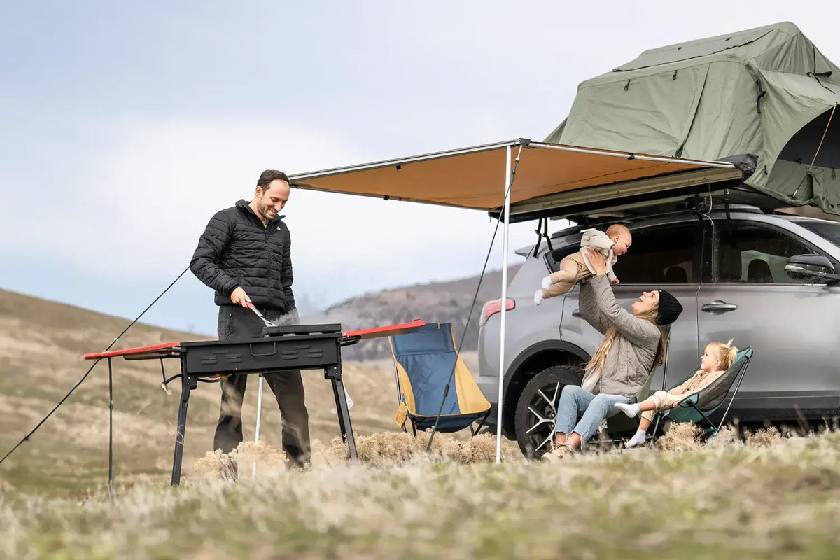A man cooks on a grill beside an SUV with a rooftop tent. A woman sits nearby holding a baby up, while a young child sits in her lap. They are outdoors on a grassy hill.