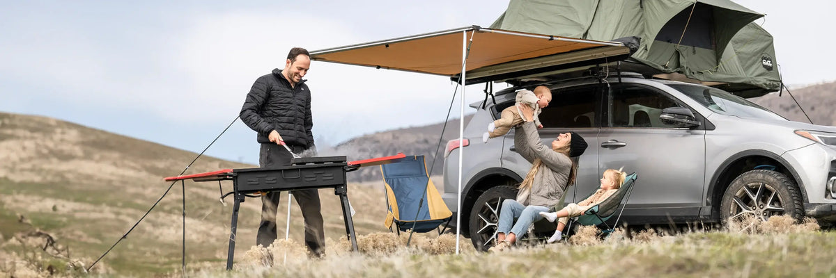 A family camps beside an SUV with a roof tent. A man grills food, while a woman sits holding a baby and a young child sits nearby. The group relaxes under an awning in a grassy, hilly landscape.