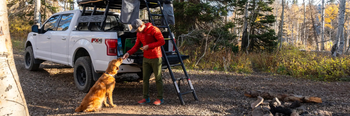 A person in a red jacket feeds a golden retriever by a white pickup truck with camping gear, parked in a forested area near a fire pit.