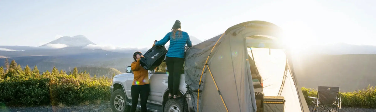 Two people set up a rooftop tent on a truck at sunrise, with mountains and greenery in the background. Camping gear and chairs are visible near the tent.