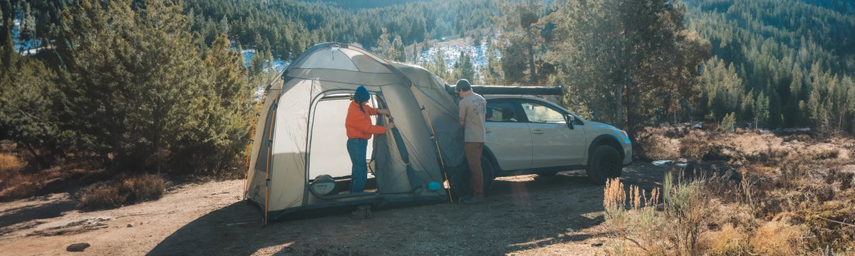Two people stand by a tent attached to a car in a forest clearing, with trees and mountains in the background, enjoying a sunny day outdoors.