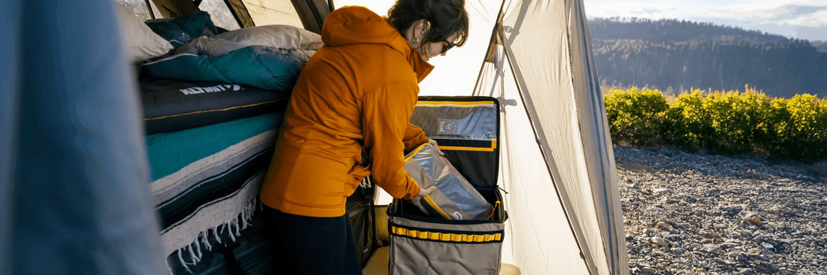 A person in an orange jacket unpacks a bag inside a tent, with bedding nearby and a scenic outdoor landscape visible through the tent opening.