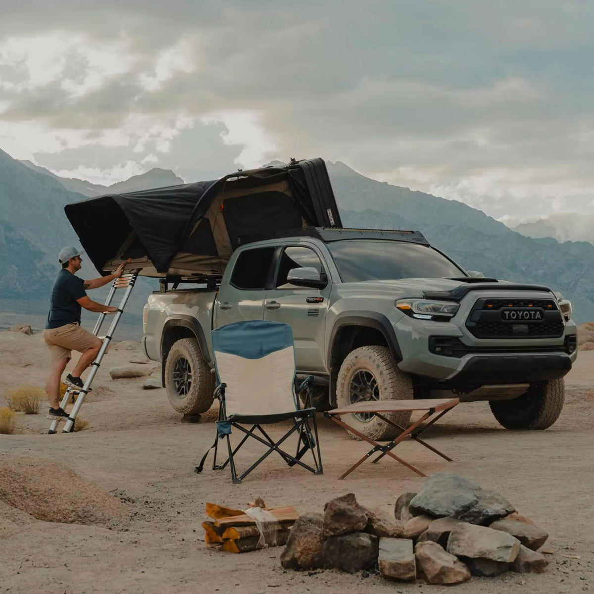 A person climbs a ladder to a rooftop tent on a Toyota truck parked on rocky terrain, with camping chairs, a fire pit, and mountains in the background under a cloudy sky.