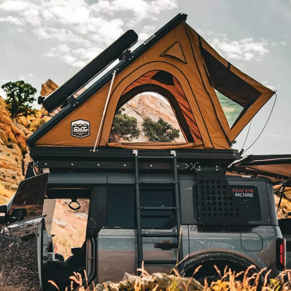 A rugged off-road vehicle with a rooftop tent is parked in a rocky, desert landscape. The tent is open, with a ladder leading up, and the vehicle doors are open, suggesting an outdoor camping adventure.