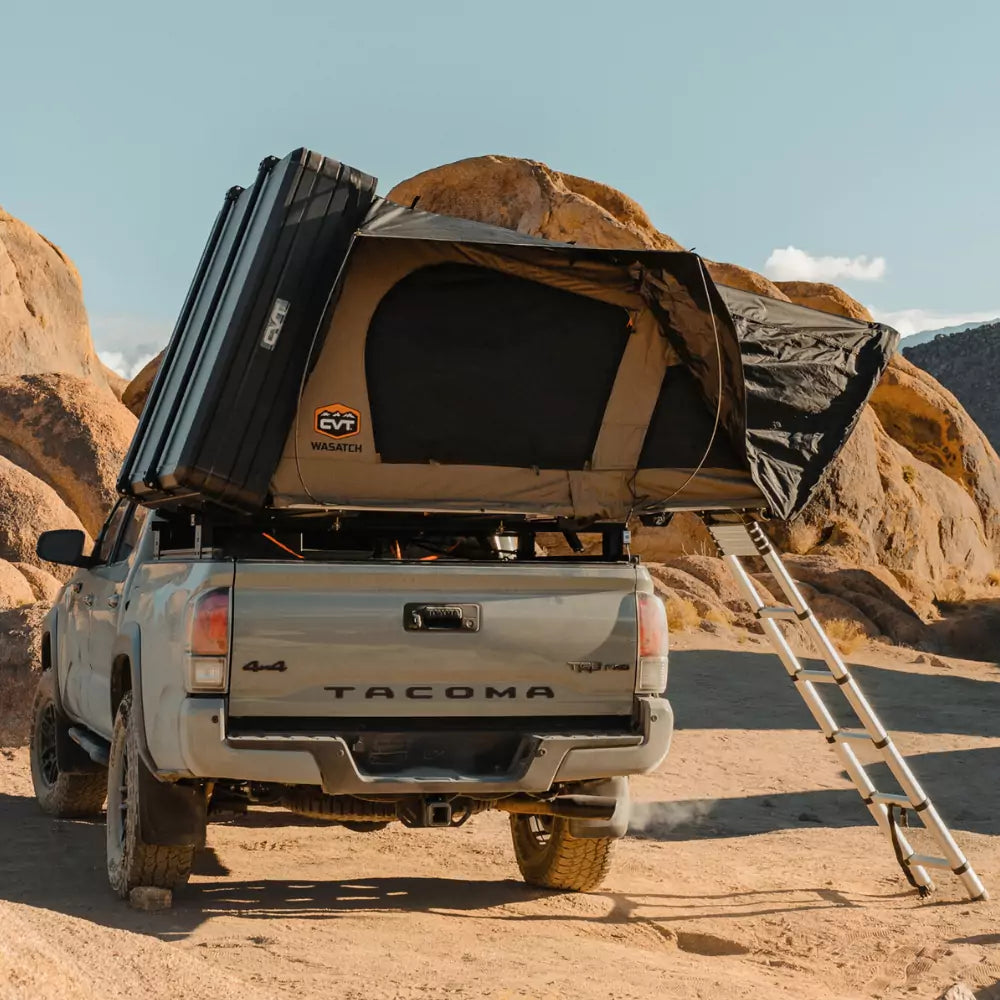 A silver pickup truck parked in a rocky desert has a rooftop tent set up with a ladder extending down to the ground.